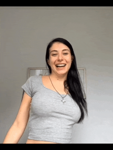 a woman wearing a grey crop top is smiling in front of a white wall