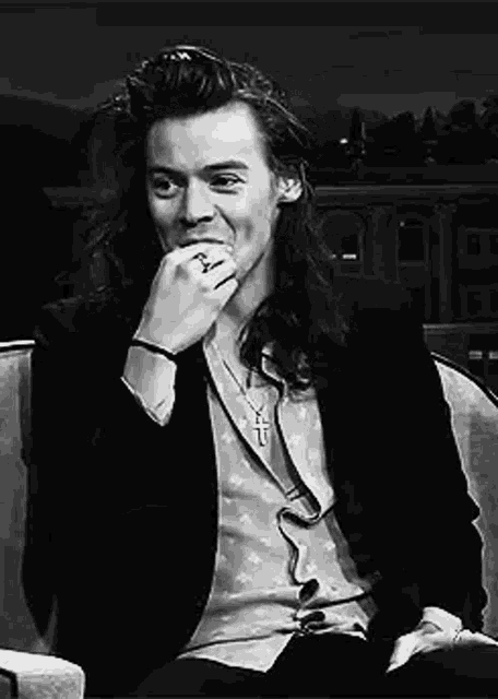 a black and white photo of a man with long hair sitting in a chair with his hand to his mouth .