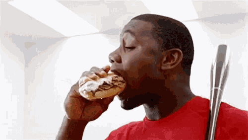 a man in a red shirt is eating a donut with a baseball bat in his hand .