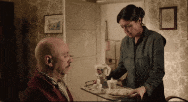 a bald man sits in a chair while a woman serves him a tray of food