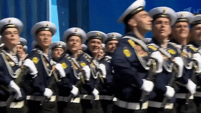 a group of men in military uniforms are marching in a line with a tv channel in the background