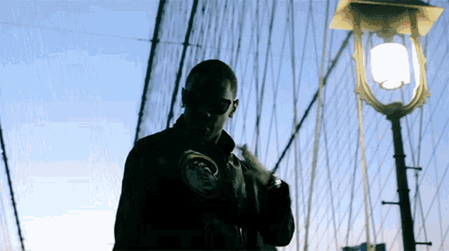 a man wearing a jacket that says ' nyc ' on it stands on a bridge