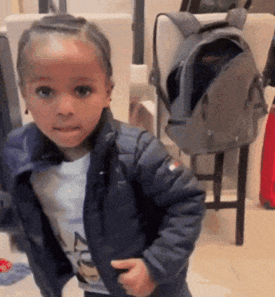 a little boy wearing a blue jacket and a white shirt is standing next to a backpack .