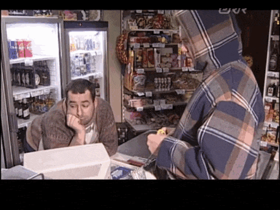a man in a plaid hoodie sits at a counter in a store while another man looks on
