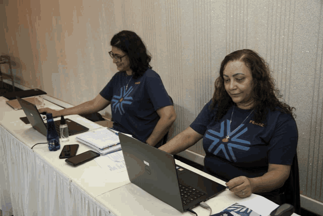 two women sitting at a desk with laptops one of whom is wearing a shirt that says ' phoenix '