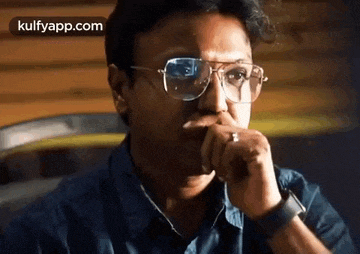 a man wearing glasses is sitting in a chair with his hand on his chin .