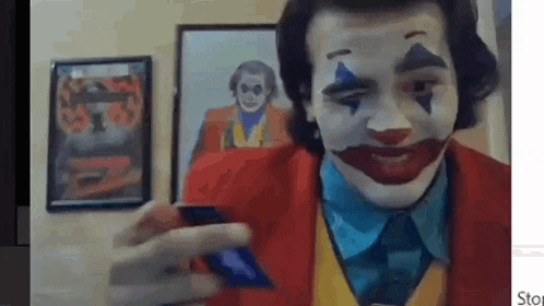 a man in a clown costume is holding a cell phone in front of a picture of the joker .