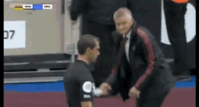 a man in a suit and tie is shaking hands with a referee on a soccer field .