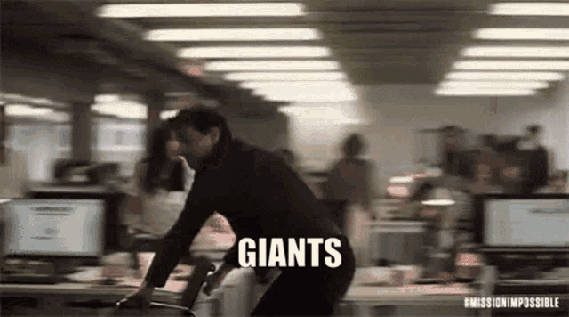 a man in a black suit is standing in an office with the word giants written on the floor .