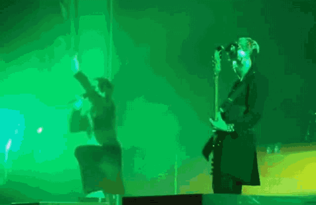 a man is playing a guitar while another man sings into a microphone on a stage in front of a green light .