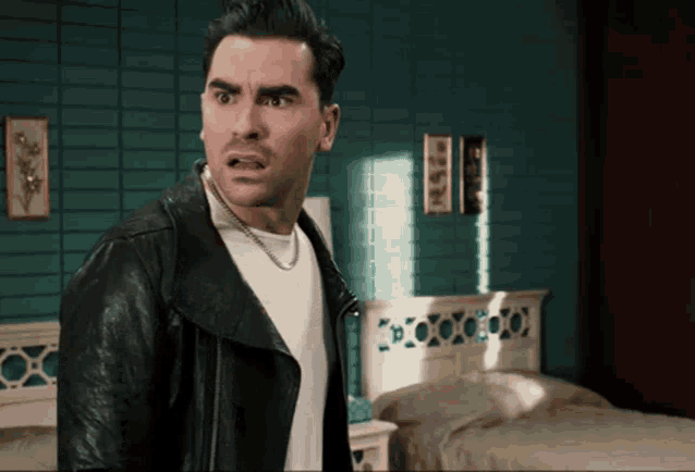 a man in a black leather jacket stands in front of a bed in a bedroom