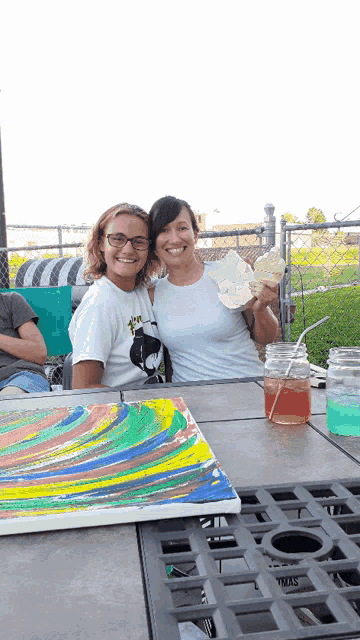 two women are sitting at a table with a painting on the table
