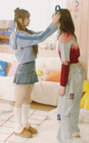 two girls are standing next to each other in a living room and one of them is putting a blue object on the other 's head