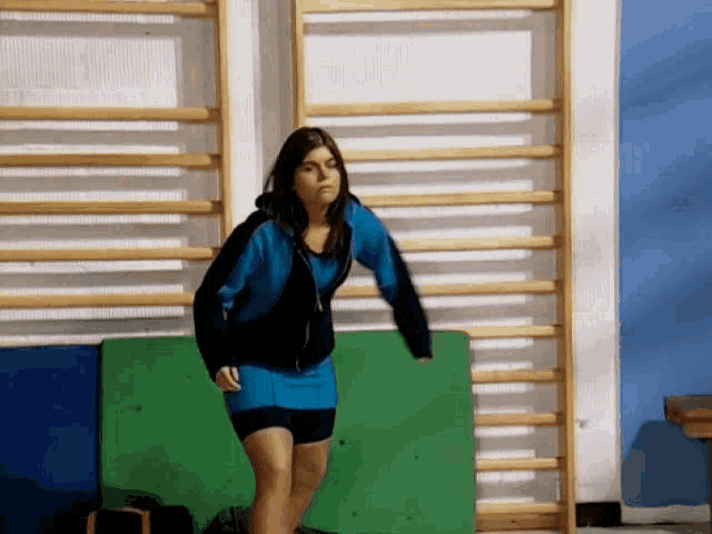a woman in a blue skirt and black jacket stands in front of a wooden wall