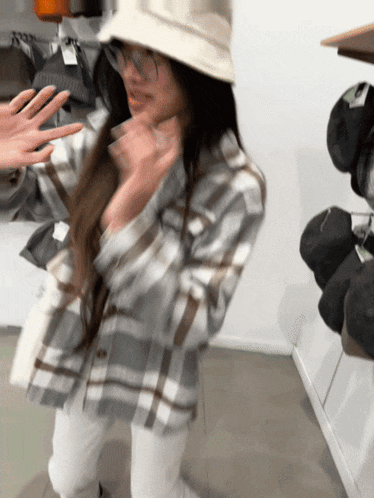 a woman wearing a plaid shirt and a bucket hat stands in front of a shelf of hats