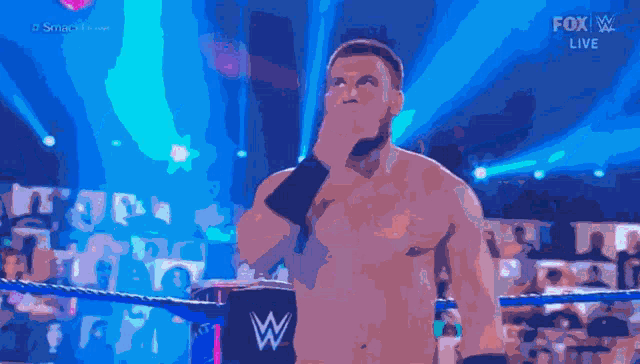 a shirtless wrestler stands in a wrestling ring with a fox live logo in the background