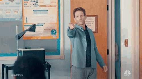 a man is pointing at the camera while standing in a classroom .