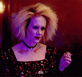 a woman with blonde hair and a choker is smoking a cigarette and says eh .