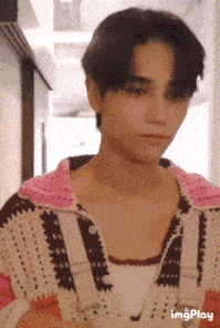 a young man wearing a pink and white sweater and a necklace is standing in a hallway .