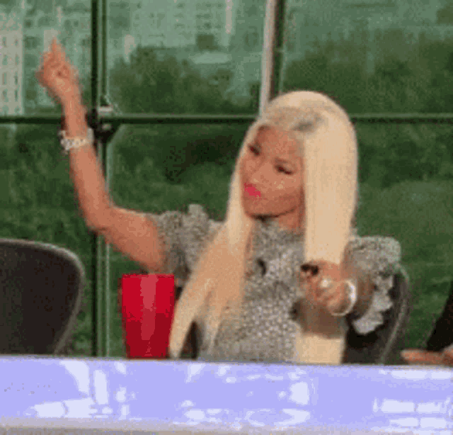 a woman with blonde hair is sitting at a table with her hand up .