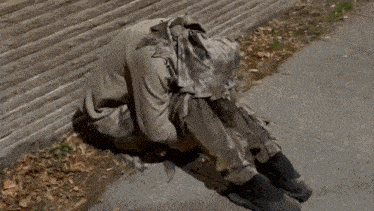 a person is kneeling down on the sidewalk with a hood on their head .