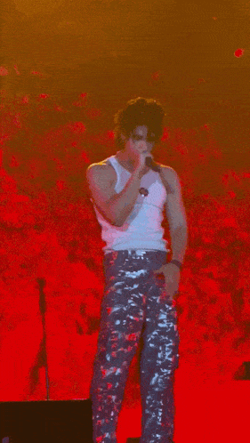 a man in a white tank top and camo pants is standing on a stage holding a microphone .