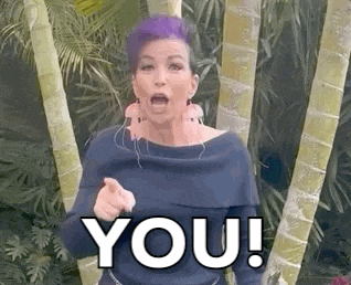 a woman with purple hair is pointing at the camera while standing in front of palm trees .