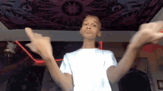 a boy in a white shirt is dancing in front of a purple tapestry on the ceiling