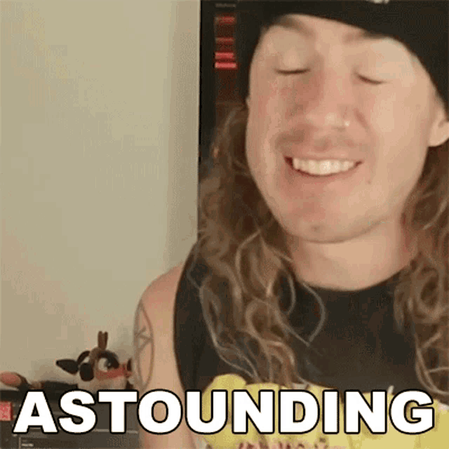 a man with long curly hair is smiling with his eyes closed and the word astounding is on the screen behind him .