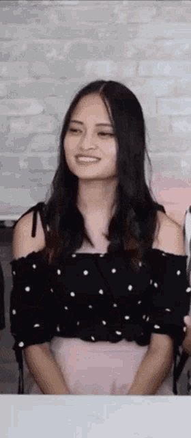 a woman in a black off the shoulder top and pink skirt is smiling and looking at the camera .