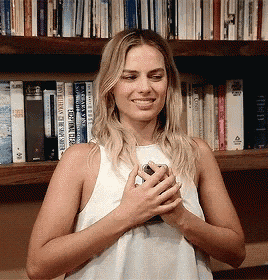 a woman in a white tank top is holding a cell phone in front of a bookshelf with books on it