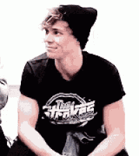 a young man wearing a black t-shirt and a black beanie is sitting on a bench .