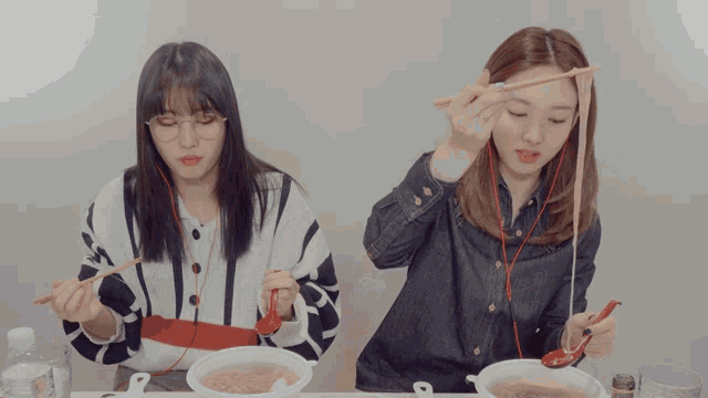 two girls are sitting at a table eating noodles with chopsticks and a twice logo on the bottom right