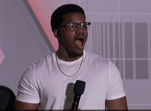 a man wearing glasses and a white shirt holds a microphone with his mouth open