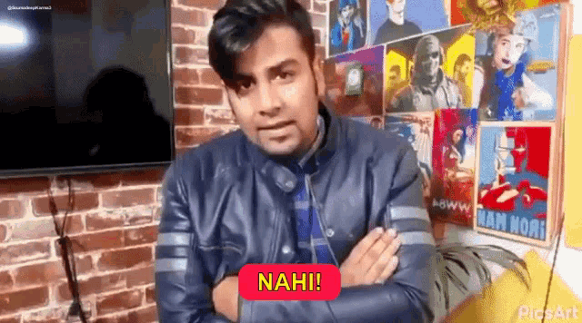 a man in a leather jacket is standing in front of a brick wall with his arms crossed and says nah !