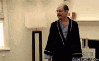 a bald man in a robe is standing in a living room with a lamp .