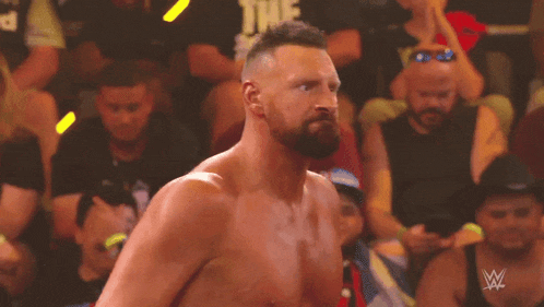 a shirtless wrestler stands in front of a crowd with a w logo on the bottom right