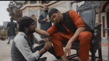 a man is sitting in a chair while another man feeds him a cigarette
