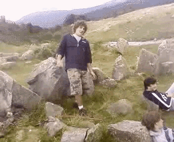 a man is standing on a rock while another man sits on a rock reading a book