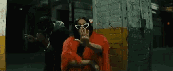 a woman wearing sunglasses and a red coat is giving the middle finger while standing next to a man .