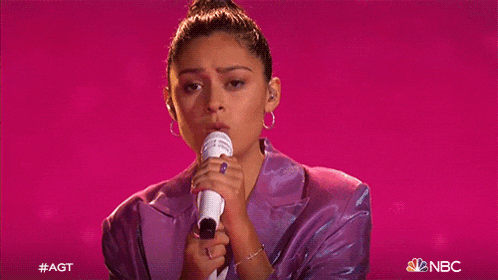 a woman in a purple suit is singing into a microphone on a pink background .