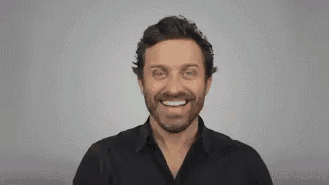 a man with a beard is smiling and pointing at the camera with his finger .