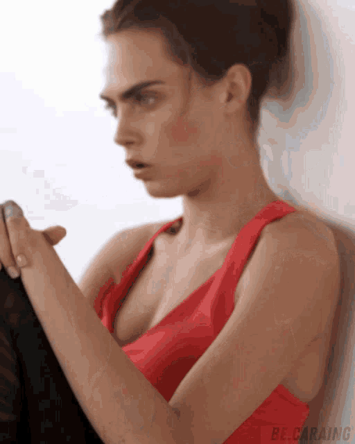 a woman in a red tank top is leaning against a wall with be.caraing written on the bottom right