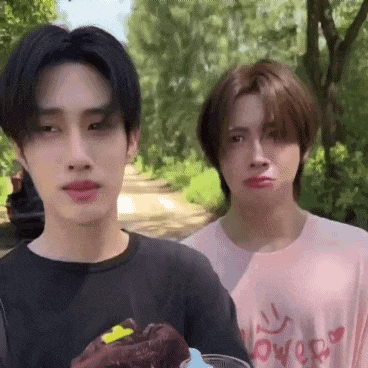 two young men are standing next to each other in a park . one of the men is holding a piece of chocolate cake .