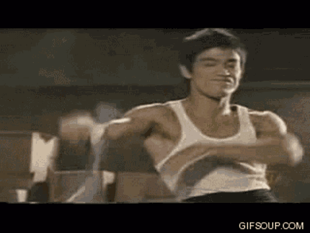 bruce lee is wearing a white tank top and boxing gloves in a boxing ring .