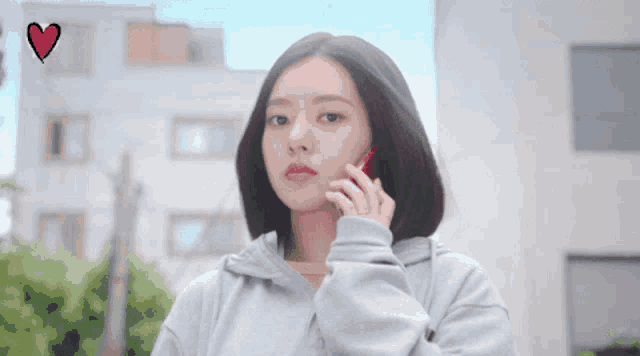a woman in a grey hoodie is talking on a cell phone with a red heart in the background