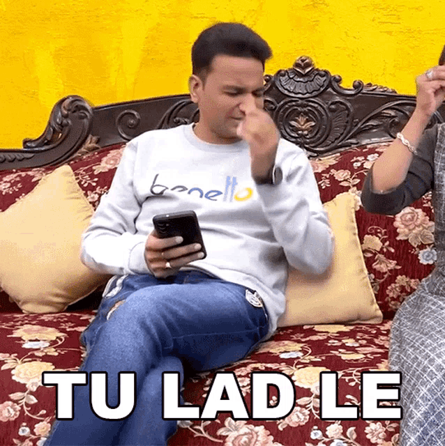a man is sitting on a couch looking at his phone and the words tu lad le are above him