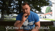 a man in a blue shirt is sitting on a bench with the words " vsiichni mi hulte curaka " on the bottom