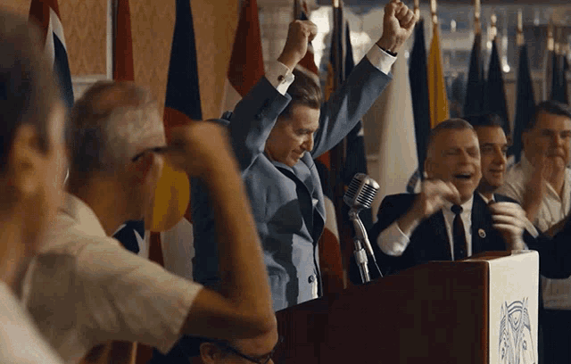 a man in a suit stands at a podium with his arms in the air