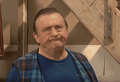 an older man wearing a blue shirt and a plaid vest is standing in front of a wooden staircase .